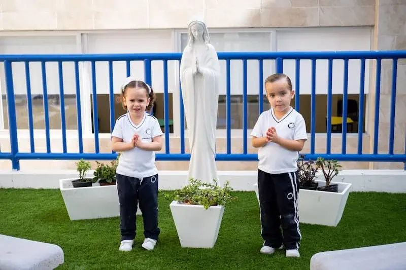 Dos estudiantes en uniforme escolar realizan ejercicio de meditación frente a estatua religiosa en zona verde del colegio